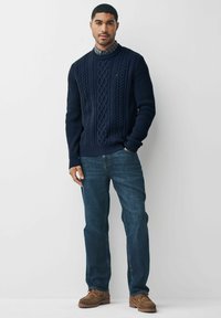 Marineblå ribbet kabelstriktrøje med rund hals, parret med lyseblå denimjeans og brune afslappede sko. Baggrunden er enkel hvid.