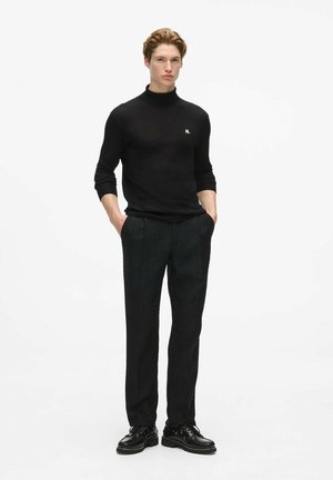 Joven de pie con las manos en los bolsillos, vestido con un jersey de cuello alto negro, pantalones oscuros y zapatos negros con cordones sobre fondo blanco.