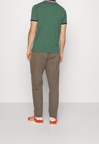 Zelené polo tričko s červenými a námořnickými pruhy, v kombinaci s hnědými kalhotami a oranžovými teniskami Adidas, viděno zezadu.