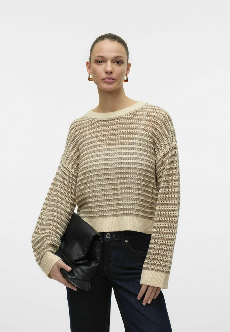 Beige gebreide trui met een losse pasvorm, voorzien van een openwerkt ontwerp en geribde manchetten. Gepaard met donkere jeans en een zwarte clutch.