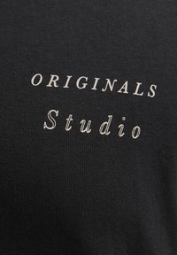 Sort stof t-shirt med den trykte tekst "ORIGINALS" i store bogstaver og "Studio" i små bogstaver, begge i en kontrasterende off-white farve.