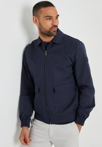 Marineblaue Bomberjacke mit Reißverschluss, zwei Fronttaschen, elastischen Bündchen und flachem Kragen. Glattes Material mit matter Oberfläche.