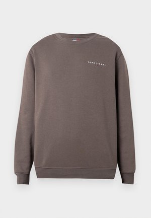 Mann trägt braunes Sweatshirt mit Rundhalsausschnitt über einem gelben Hemd mit hochgekrempelten Ärmeln, kombiniert mit dunkelblauer Hose und braunem Gürtel, Hände in den Taschen.