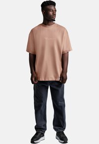 Maglietta rosa chiaro con scollo a girocollo e maniche corte, con testo bianco ricamato. Abbinata a jeans di denim scuro e sneakers nere.