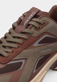 Κοντινή εικόνα καφέ αθλητικού παπουτσιού Calvin Klein με υφασμένες κορδέλες, πάνελς από σουέντ και δίχτυ, καθώς και με το λογότυπο στη γλώσσα και στο πλάι.