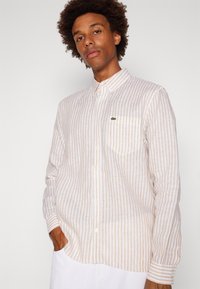 Camicia button-up a righe di un beige chiaro con colletto. Realizzata in tessuto traspirante, presenta una tasca sul petto e un dettaglio logo sottile.