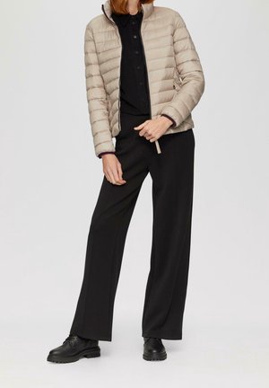 Beige gewatteerde pufferjas met een opstaande kraag, gecombineerd met zwarte wide-leg broeken en zwarte veterschoenen. Gladde textuur, minimale details.