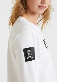 Vit långärmad t-shirt i bomull med en svart fyrkantig logotyp på vänster ärm med texten "OFF THE WALL" och geometriska accenter på bröstet.