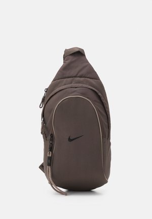 Brązowy plecak Nike na jedno ramię z przednią kieszenią na zamek i regulowanym paskiem na białym tle.