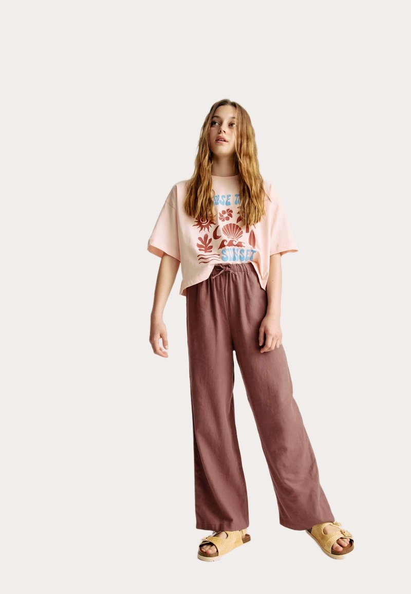 Lichtroze cropped T-shirt met een grafisch ontwerp, gecombineerd met losse bruine linnenbroek en beige slip-on sandalen.