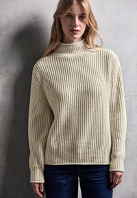 Maglione a collo alto a coste crema con maniche lunghe, caratterizzato da un motivo di maglia testurizzato e una vestibilità ampia, abbinato a jeans blu scuro.