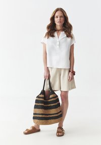Hvid kortærmet skjorte med knaplukning, beige linned shorts, og en stor stribet vævet tote taske i sort og tan, parret med brune sandaler.