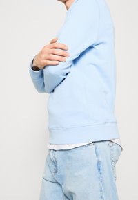 Sudadera azul claro con cuello redondo y puños acanalados. Llevada sobre una camisa blanca, combinada con jeans de mezclilla de lavado claro. Textura suave.