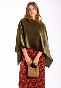Poncho de satén verde oliva, con diseño drapeado y una textura suave. Combinado con un vestido floral en tono burdeos y un pequeño bolso de mimbre beige.
