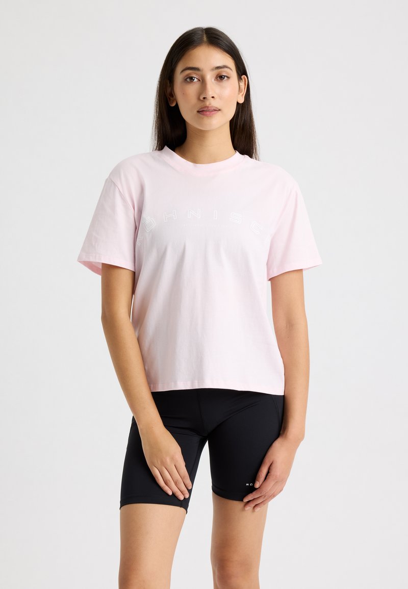 Camiseta de algodón rosa claro con cuello redondo y mangas cortas, con texto blanco en un diseño curvado. Combinada con pantalones cortos de ciclismo negros.