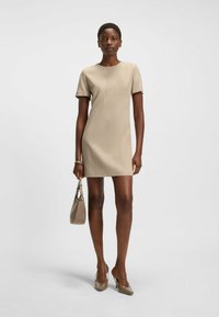 Beige klänning med kort ärm och rak silhuett. Slät textur, minimalistisk design, bärs med en matchande handväska och spetsiga klackar.
