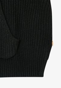 Μαύρη ριγέ πλεκτή μανσέτα που έχει μπροστινή τσέπη και μια μικρή μπεζ υφασμάτινη ετικέτα στο πλάι.