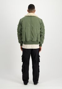 Veste bomber vert olive avec un col en fausse fourrure blanche, des poignets côtelés et une ceinture ; portée sur une chemise beige clair et un pantalon cargo noir.