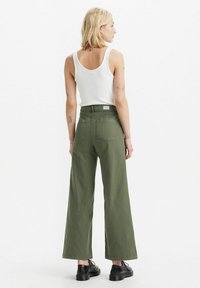 Grüne weit geschnittene Baumwollhose mit hoher Taille, Gesäßtaschen und leicht strukturiertem Finish, kombiniert mit einem weißen Tanktop.