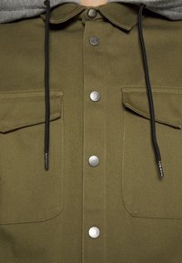 Primer plano de una chaqueta verde oliva con bolsillos de solapa, botones a presión plateados, cuello y cordones negros que provienen de una capucha gris.