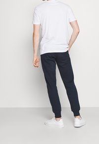 Mies, który ma na sobie białą koszulkę z krótkim rękawem, czarne dresy z tylną kieszenią oraz białe sneakersy na jednolitym jasnym tle.