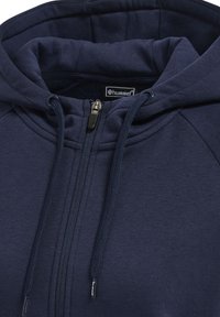 Felpa con zip di colore navy in tessuto morbido, dotata di cappuccio regolabile con cordino, zipper visibile e fodera interna a contrasto. Etichetta del marchio esposta all'interno.