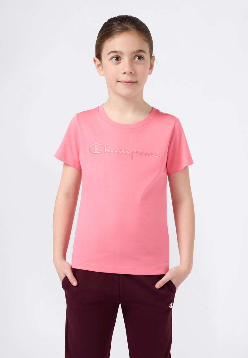 Νεαρός άνθρωπος στέκεται με τα χέρια στις τσέπες, φορώντας ένα ροζ μπλουζάκι Champion και σκούρα μαρούλι παντελόνι μπροστά από έναν απλό φόντο.