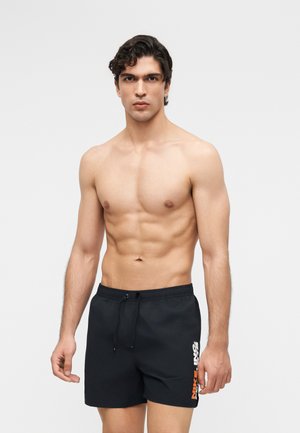 Άνδρας χωρίς πουκάμισο με κοντά σγουρά μαλλιά στέκεται στραμμένος προς τα εμπρός, φορώντας μαύρα μαγιό Nike με το λογότυπο στο αριστερό μηρό σε απλό φόντο.