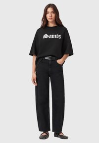 Zwarte oversized t-shirt met "Saints" in witte schrift, gecombineerd met hoge taille zwarte wijde jeans en studded zwarte platte schoenen.