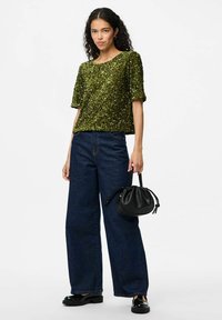 Top corto a maniche corte verde con paillettes abbinato a jeans in denim blu scuro a gamba larga. Una borsa rotonda nera è tenuta in una mano, accentuando l'outfit.