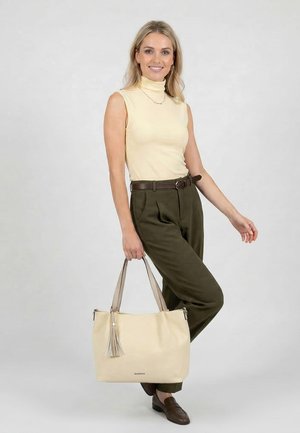 Kobieta stojąca i uśmiechająca się, ubrana w bezrękawnik beżowy, oliwkowe spodnie z paskiem, trzyma dużą beżową torebkę z frędzlem, brązowe mokasyny.