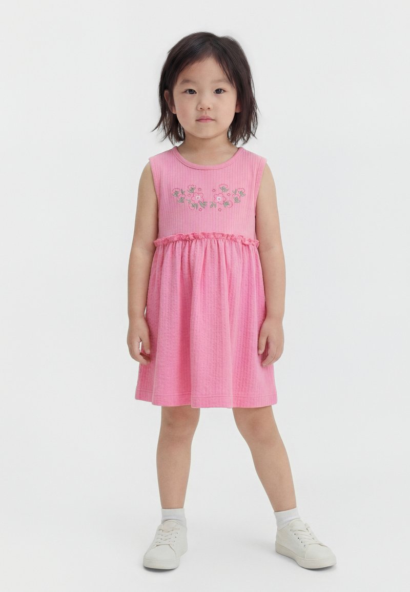 Νεαρό κορίτσι στέκεται με τα χέρια δίπλα στο σώμα, φορώντας ροζ φόρεμα χωρίς μανίκια με κεντήματα λουλουδιών και άσπρα αθλητικά παπούτσια σε απλό φόντο.