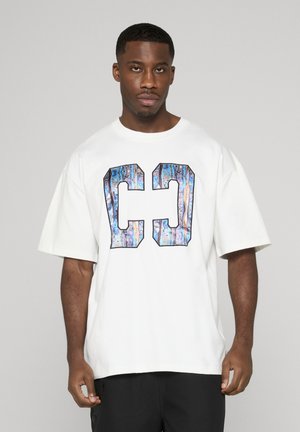 Mann mit kurzen Haaren trägt ein weißes übergroßes T-Shirt mit großem blau-lila gemustertem "CC"-Design, steht vor einem schlichten Hintergrund.