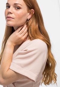 Helles, rosa, strukturiertes Kleidungsstück mit kurzen Ärmeln; das Modell hat lange, wellige, rote Haare und trägt einen kleinen, gebogenen Ohrring.