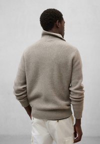 Beige gerippter Strickpullover mit umgeschlagenem Kragen, langen Ärmeln und lässiger Passform, getragen über weißen Hosen.