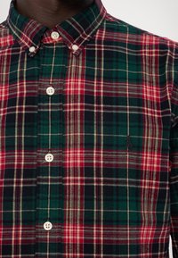 Camicia a scacchi con bottoni, caratterizzata da un mix di colori rosso, verde e blu navy, con colletto classico e tre bottoni bianchi sulla patta.