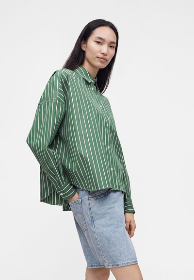 Femme aux longs cheveux foncés portant une chemise à rayures verticales vertes et blanches boutonnée, et un short en jean bleu clair, debout, une main dans la poche.