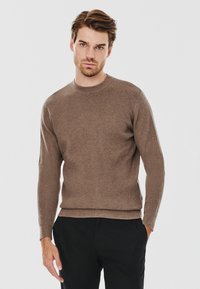 Brązowy, prążkowany sweter z okrągłym dekoltem i długimi rękawami. Materiał wydaje się miękki, o dopasowanym kroju. Nosi się go na czarnych spodniach.