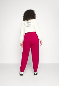 Person med lockigt hår som bär en vit kortärmad sweatshirt med texten "Adidas Athletic League" och magentaröda träningsbyxor och vänder sig bort från en vit bakgrund.