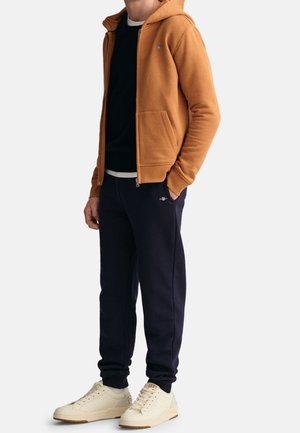 Man die een bruine hoodie met rits draagt, zwarte trainingsbroek en crèmekleurige sneakers, staand met één hand in zijn zak.