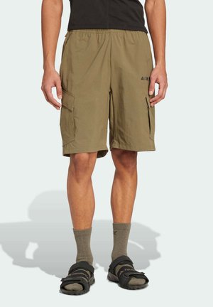 Mann trägt olivgrüne Cargoshorts, graue Socken und schwarze Sandalen, steht vor einem schlichten hellen Hintergrund.