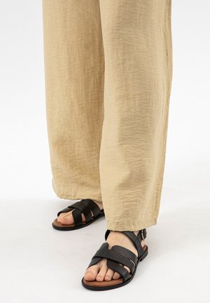 Feet wearing black leather sandals with wide straps and beige wide-leg pants against a white background.