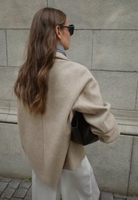 Bež suurendatud villase segu jakk struktuursete õlgadega, koos musta käekotiga. Pikad, lainelised juuksed ja hall sall on nähtavad.