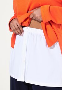 Oranžová přehozená mikina přes bílou košili s knoflíky, s elastickým páskem, který vystavuje pás a dolní část košile.