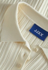Kremasta rebrasta polo majica s tri gumba, mekim teksturom i plavom oznakom "JJXX" blizu ovratnika.