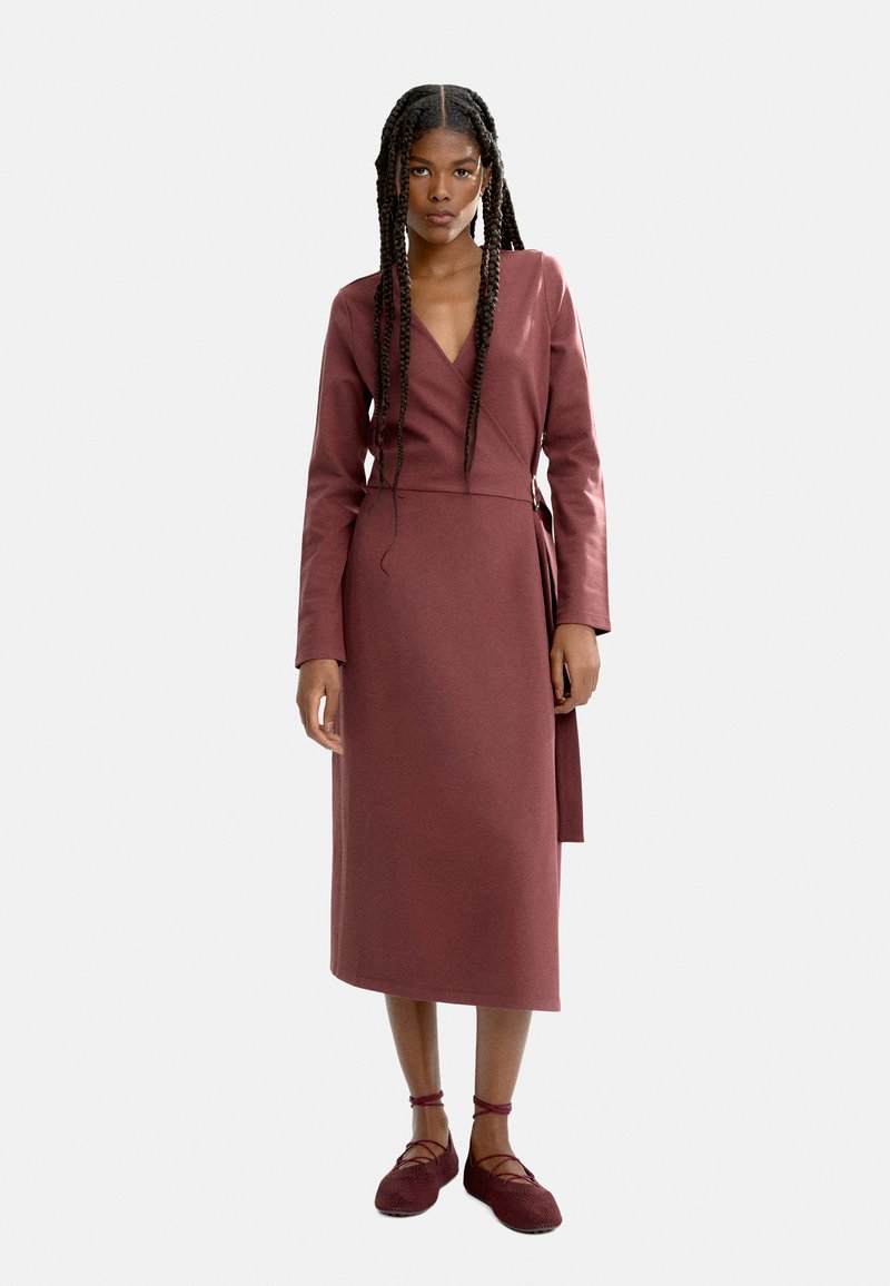 Bordowa sukienka kopertowa z długimi rękawami i głębokim dekoltem w serek, z wiązaniem w talii. Modelka nosi pasujące bordowe baleriny.