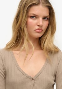 Beżowy, prążkowany top z długim rękawem, z dekoltem w kształcie litery V, z zapięciem na guziki i teksturowanym wykończeniem. Miękka tkanina z delikatnym połyskiem.