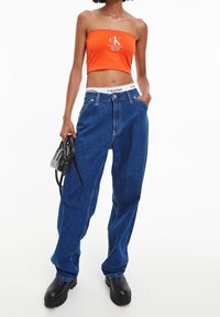 Orange crop top med vit logotyp, kombinerad med löst sittande blå denimjeans med en Calvin Klein-märkt midja. Svart handväska och kraftiga stövlar.