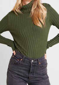 Pull à col roulé côtelé vert foncé à manches longues, coupe ajustée et col montant, associé à un jean noir taille haute avec des éléments en cuivre.
