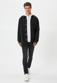 Mustaparantaisessa paidassa on napit, ja se on puettu valkoisen hupparin päälle. Slim fit -mustat farkut ja valkoiset lenkkarit täydentävät asun. Pintakuvio on sileä.